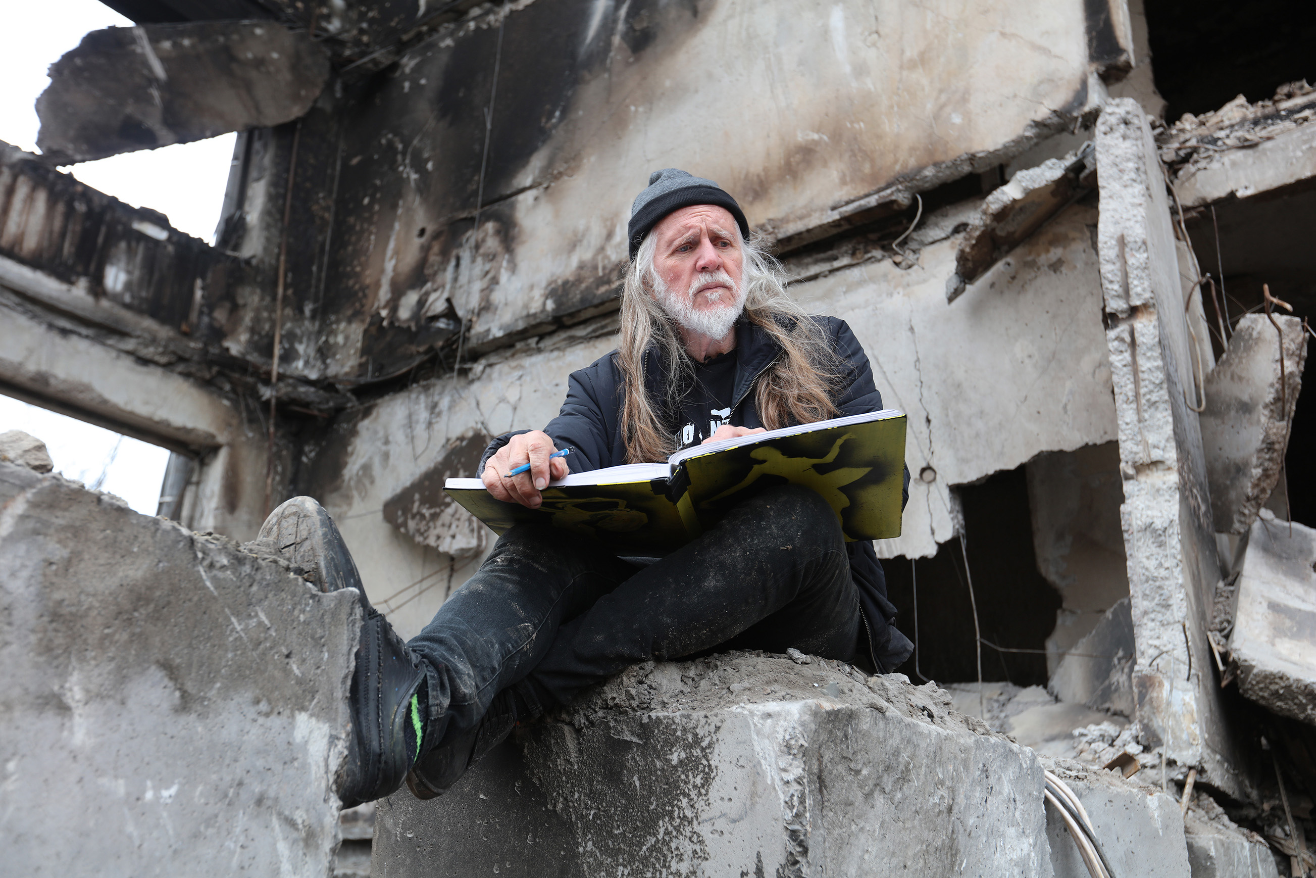 A photograph of artist George Gittoes sitting amongst the rubble of a bombed out building with a sketchpad open on his lap.